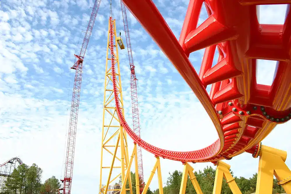 Bright red roller coaster track curving around a tall yellow support tower against a blue sky.