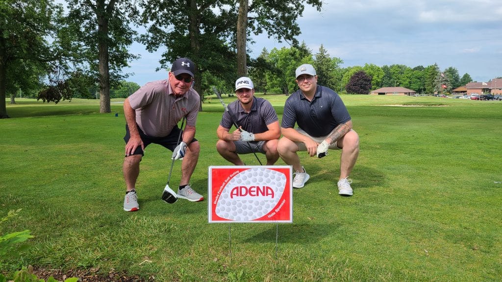 Three men crouching on a golf course holding golf clubs behind a sign that reads ‘ADENA’ with ‘Skywood Robotics Golf Outing’ and ‘Hole Sponsor’ along the sides.