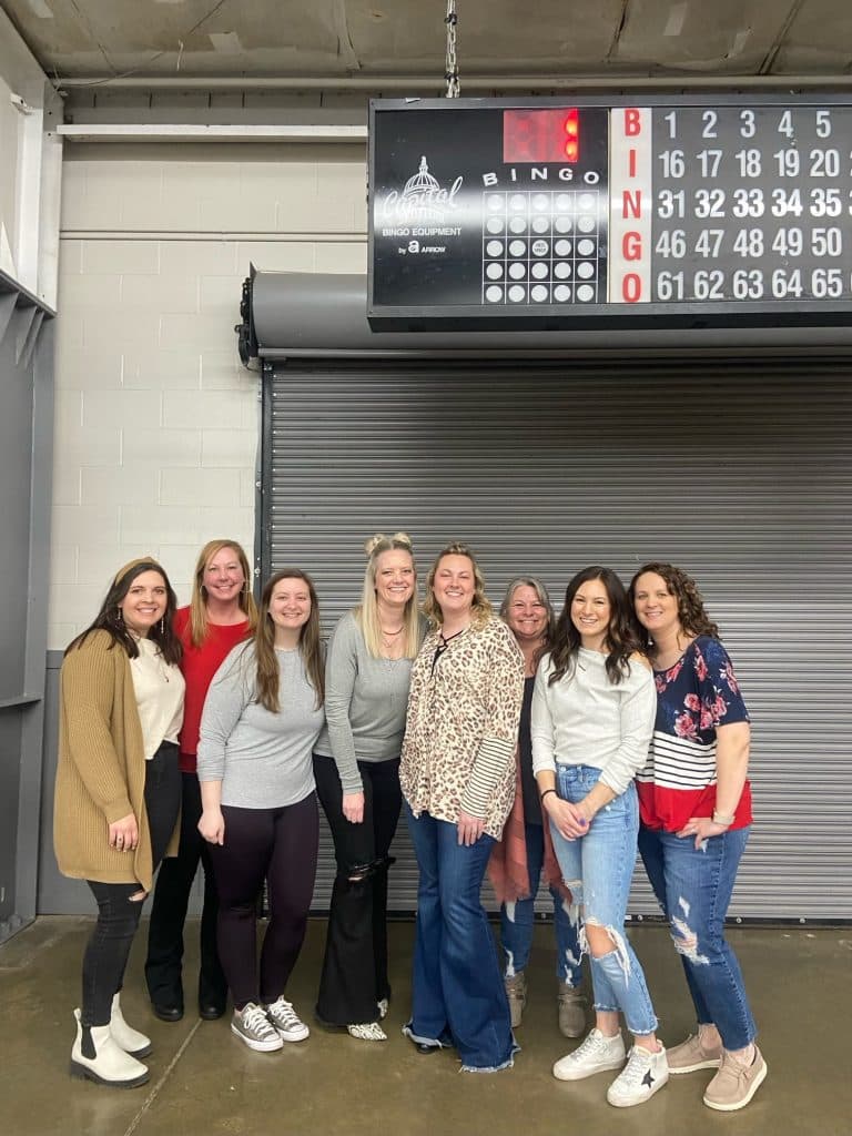 Group of women standing together in front of a closed metal roll-up door beneath a bingo scoreboard.
