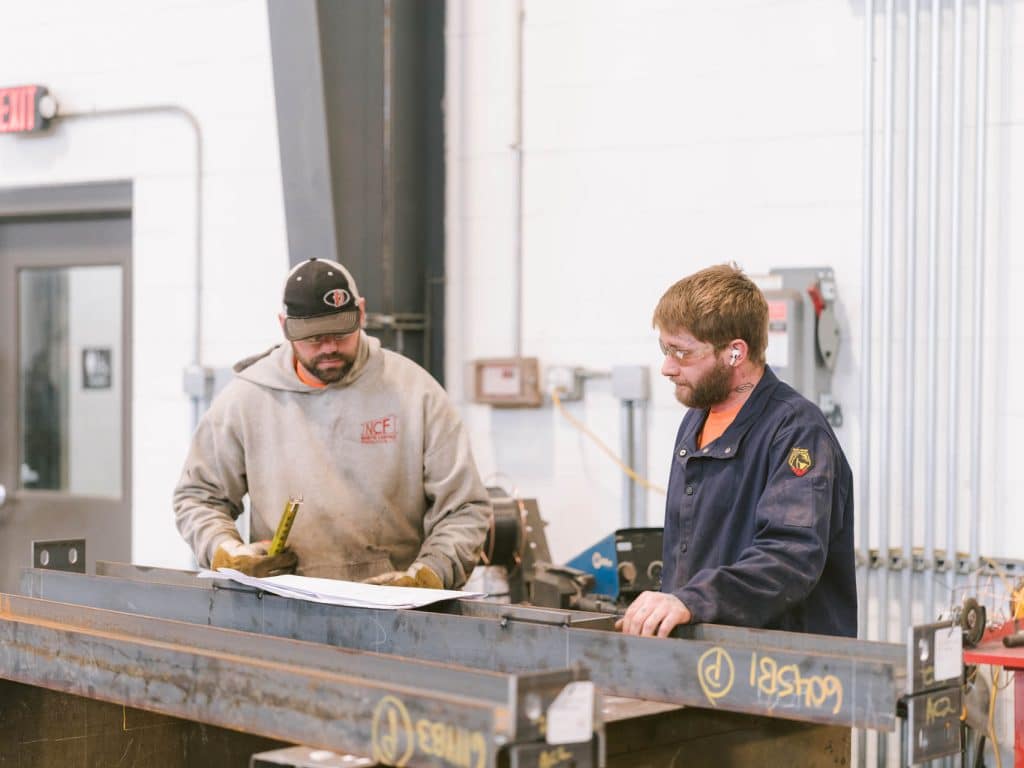 Two workers in safety glasses reviewing plans beside a large steel beam inside an industrial fabrication shop.