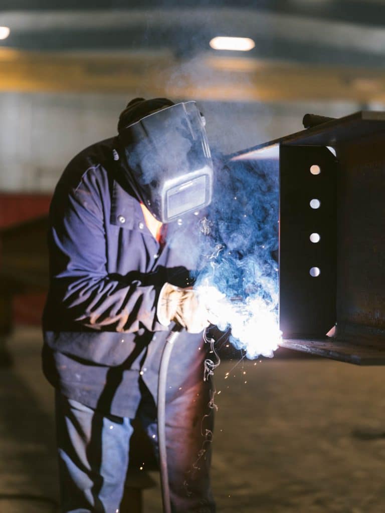 A worker wearing a welding mask and gloves welding a metal frame, with sparks and bright light visible at the weld point.