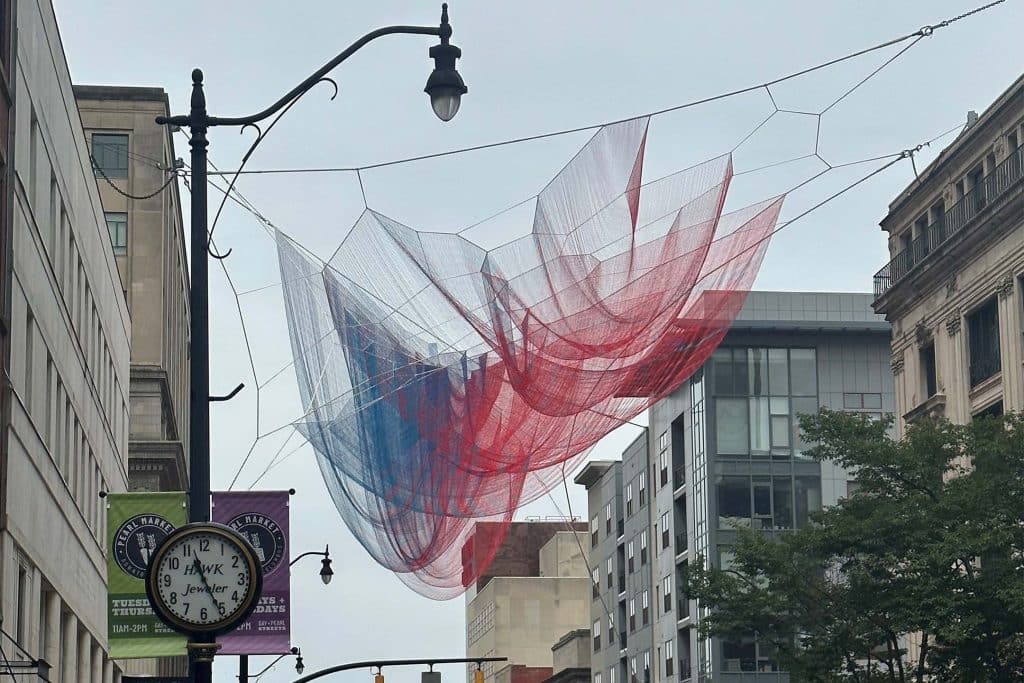 A large, billowing red, white, and blue mesh art installation suspended above a city street between buildings, with a vintage street clock and lamppost in the foreground.